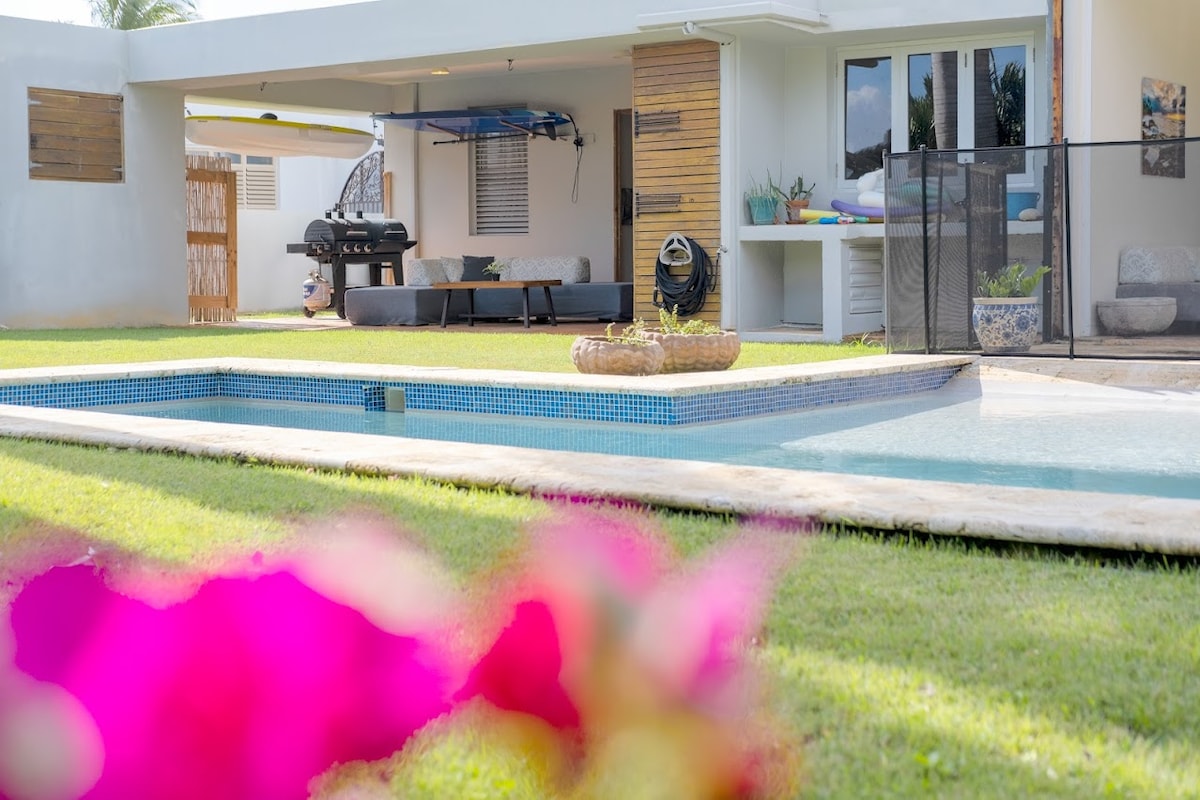 A swimming pool is situated in the foreground, surrounded by well-maintained grass. In the background, a spacious patio area features modern furnishings and potted plants. A barbeque grill is also visible, enhancing outdoor dining possibilities.