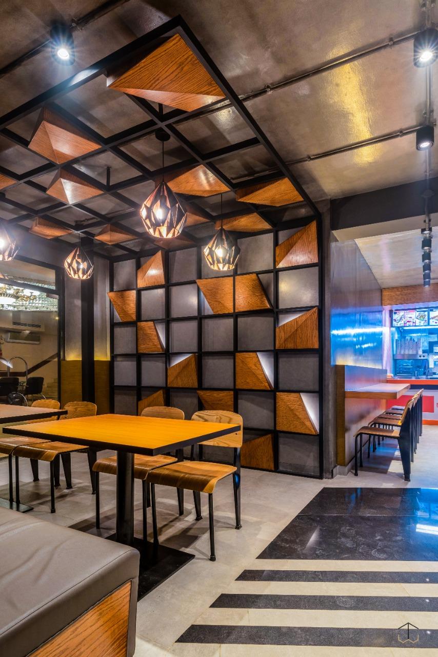 A modern dining area features a geometric wooden accent wall, showcasing varying shades of brown. Sleek pendant lights are suspended from the ceiling, illuminating the space. Simple wooden tables and chairs provide a relaxed seating arrangement, complemented by a dark floor design.