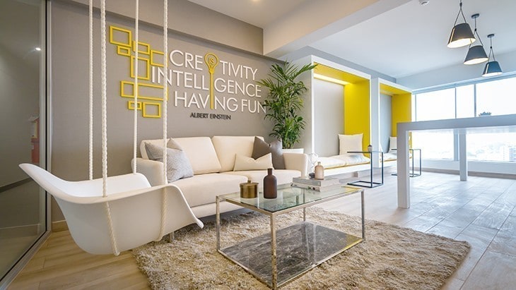 A modern living room is featured, highlighting a white sofa and a glass coffee table atop a soft, textured rug. Bright yellow accents decorate the space, along with a motivational wall display. Natural light enters through large windows, enhancing the airy atmosphere.