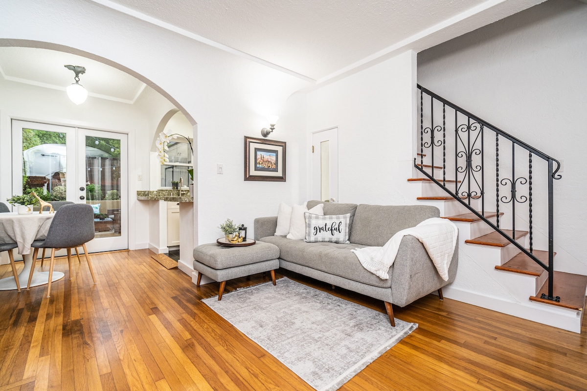 The living area is presented with a light grey sectional sofa and a plush area rug, enhancing the hardwood floor. Natural light fills the space through large glass doors, leading to a patio. A staircase with an elegant wrought-iron railing is visible, adding character to the room.