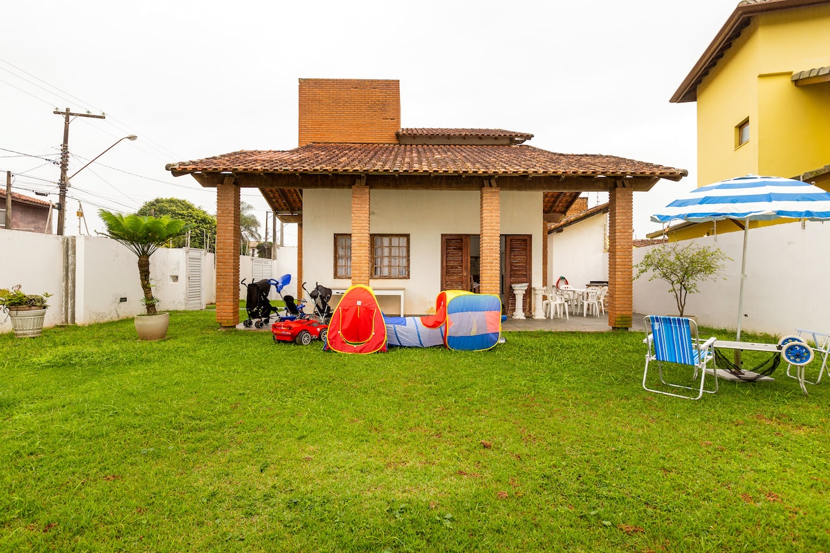 A spacious outdoor area is featured, surrounded by a lush green lawn. Colorful play tents and a red ride-on toy provide engaging options for children. Lawn chairs and a striped umbrella offer shaded seating, while the wooden patio showcases a rustic architecture under a tiled roof.