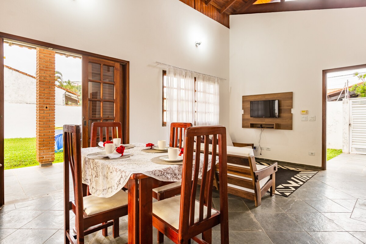 A dining area is featured with a wooden table set for four, adorned with white plates and utensils. Large windows allow natural light into the space, while a comfortable seating area is visible nearby with a TV mounted on the wall.