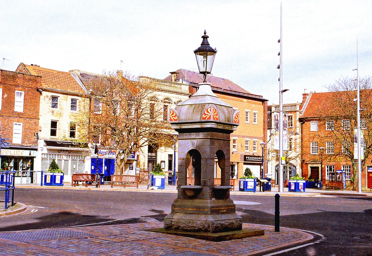 A historic water fountain is situated in the center of a quaint square, surrounded by charming buildings with a mix of architectural styles. Outdoor seating areas highlight the vibrant community space, while lampposts and planters enhance the atmosphere. Clear skies and a serene ambiance invite leisurely strolls.