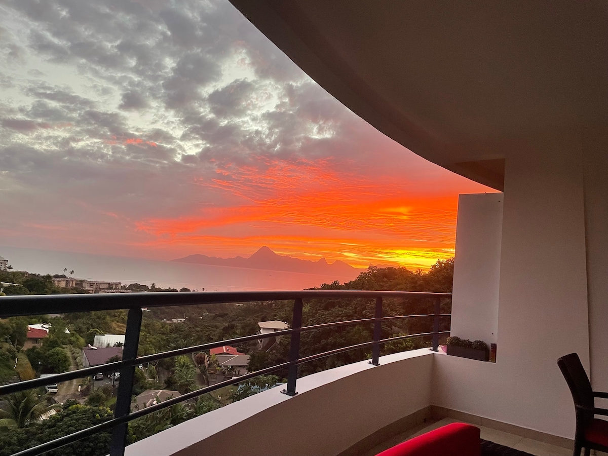 A sunset view from a spacious balcony showcases vibrant colors of orange and pink blending into the sky. The silhouette of lush greenery and distant mountains provide a serene backdrop, while the railing of the terrace frames the picturesque scene.