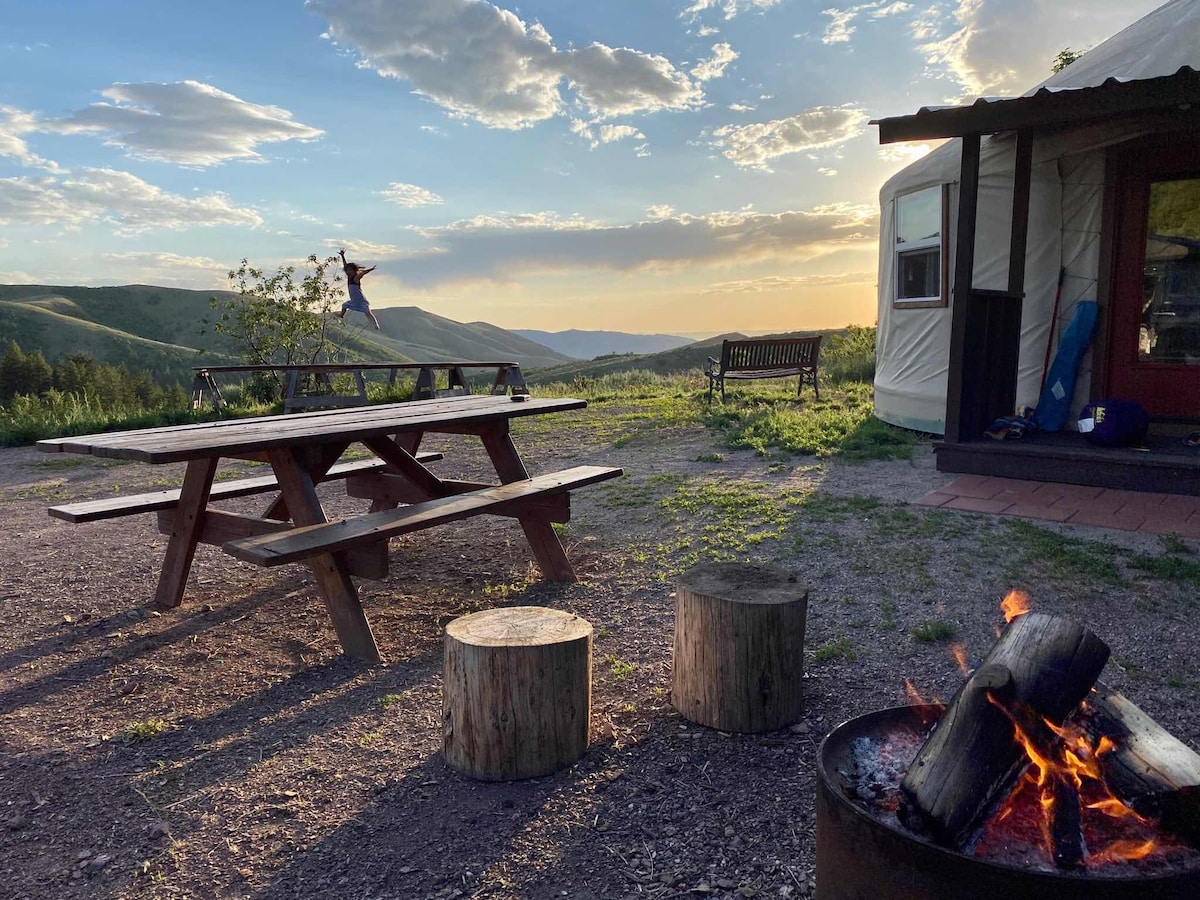 A picnic area is featured with a wooden table set against a backdrop of rolling hills and a colorful sunset. A fire pit with a small fire is visible nearby, accompanied by wooden stumps for seating. The scene captures the tranquility and natural beauty of the surroundings.
