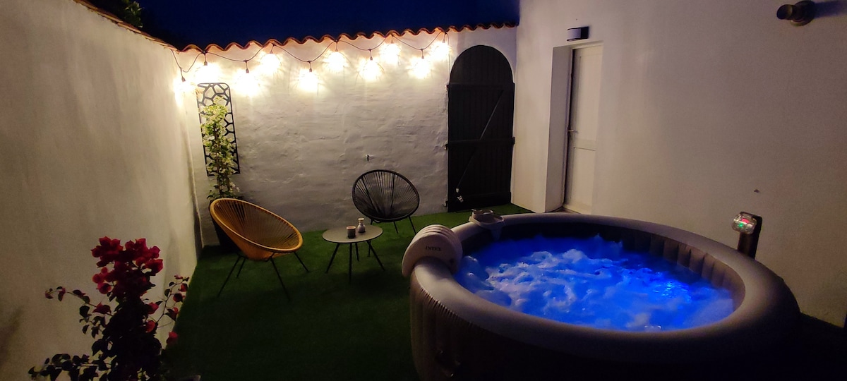 A cozy patio area features a jacuzzi with illuminated blue water, surrounded by greenery and soft lighting. A pair of lounge chairs and a small table are positioned nearby, enhancing the relaxing atmosphere. A wooden door provides access to the space.