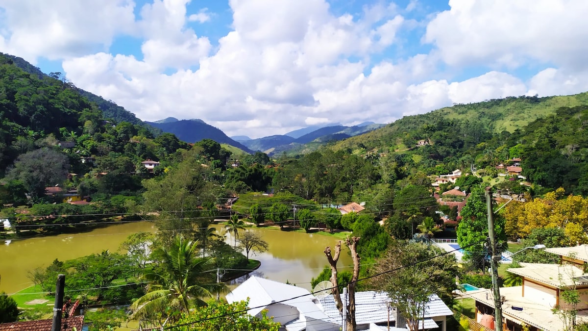 A panoramic view of the surrounding mountains and lush greenery is presented, with a serene lake at the forefront. Houses with varied architecture can be seen nestled among the trees, creating a harmonious blend of nature and residential spaces under a partly cloudy sky.