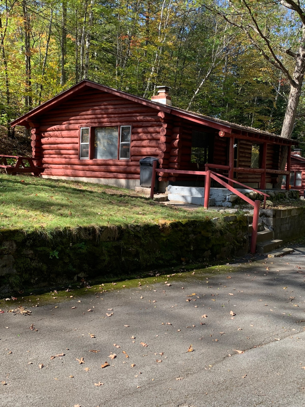 A rustic cabin is nestled among trees, featuring a distinctive red log exterior. A stone foundation supports the structure, and a wooden deck extends in front. Windows allow natural light to enter, while the surrounding foliage exhibits autumn colors.