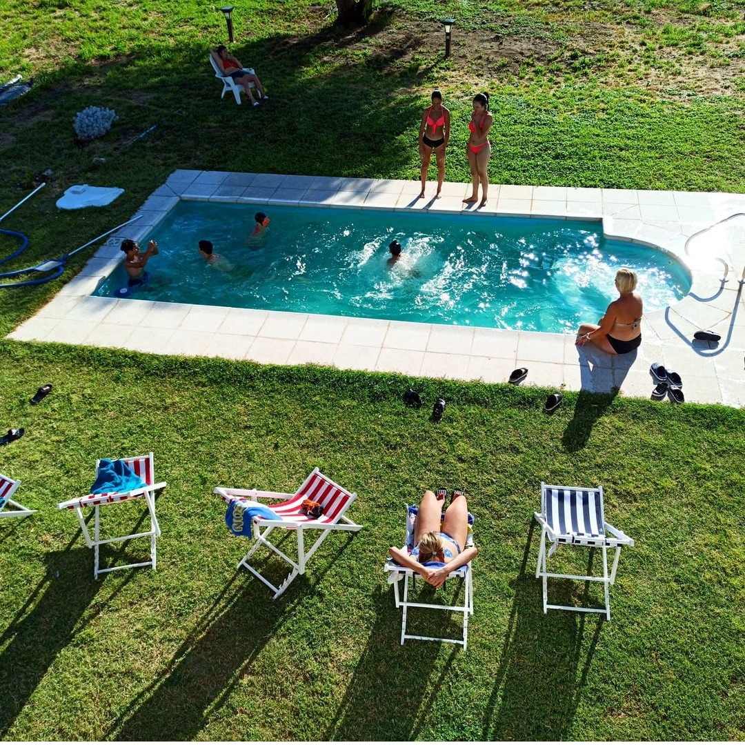 A spacious pool area is visible, surrounded by well-kept green grass. Several guests enjoy the refreshing water in the pool, while others relax on striped lounge chairs nearby. Sunlight casts gentle shadows across the scene, enhancing the calm outdoor atmosphere.