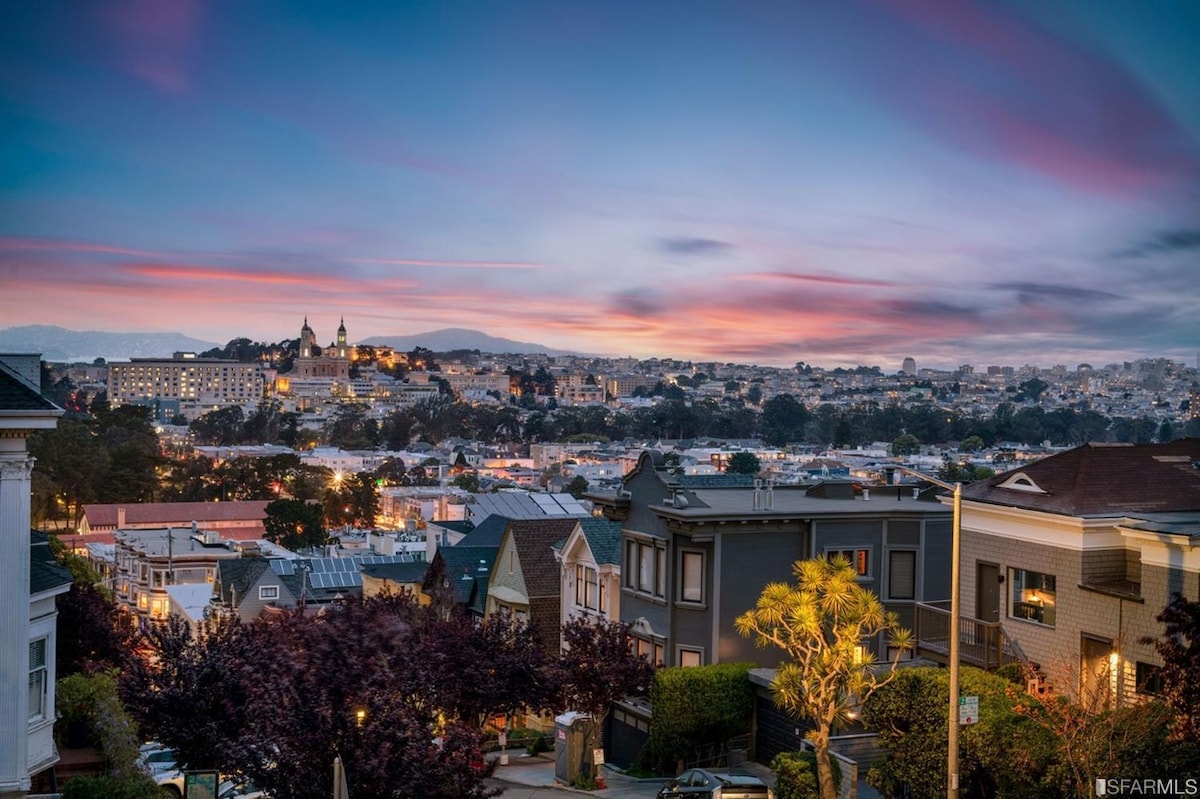 A panoramic view of San Francisco showcases the cityscape during sunset, with colorful skies transitioning from blue to pink. The foreground features residential buildings and trees, while the hills in the background highlight the iconic structures above.