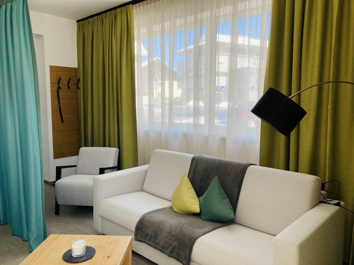 A light-filled living area features a comfortable sofa adorned with colorful cushions and a partially draped throw. Sheer curtains allow natural light to fill the space, while a side chair and a wooden table complement the inviting atmosphere.