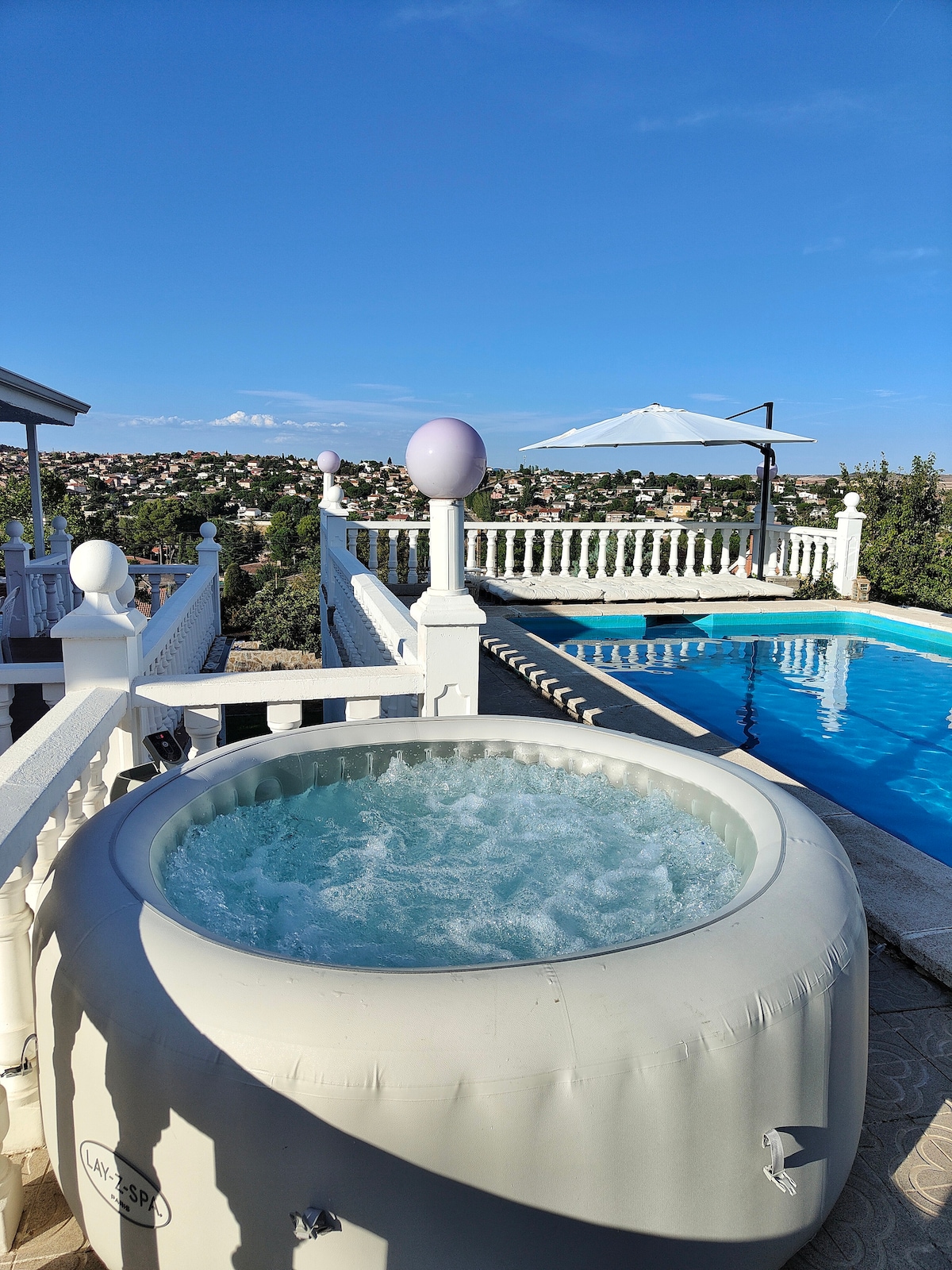 A spacious jacuzzi sits beside an inviting pool, offering a view of the surrounding landscape. The area is enhanced by a sun umbrella and elegant railings, providing a serene space for relaxation. Clear skies create a bright atmosphere.