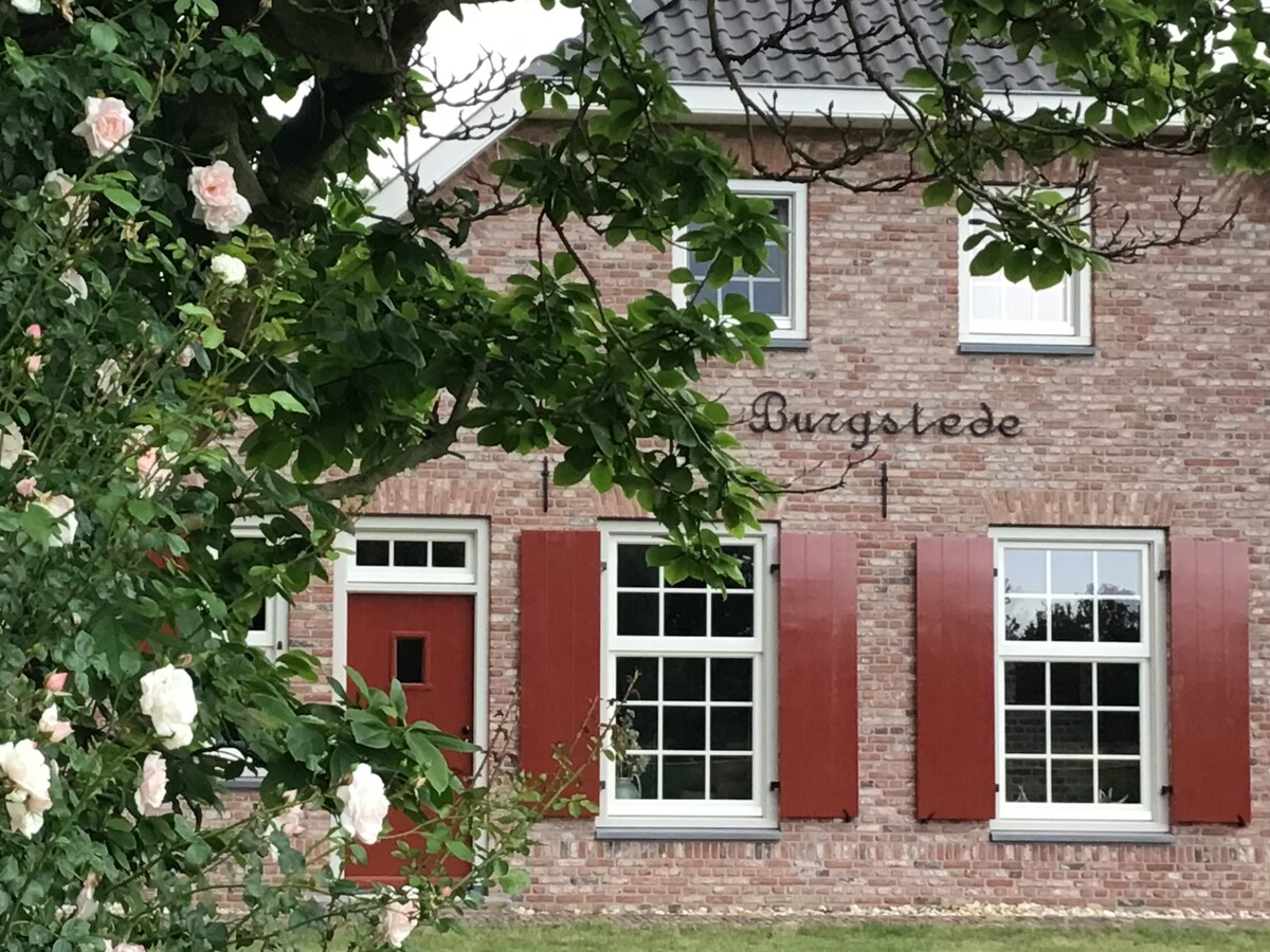 A charming brick exterior of the farmhouse is framed by a lush rose bush. The building features multiple large windows with red shutters, complemented by a distinctive red door. The greenery of surrounding trees adds to the natural setting.
