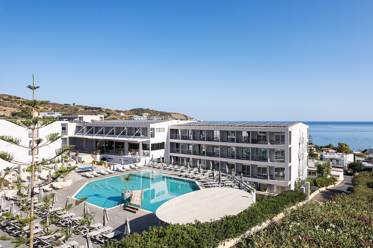 An aerial view of Atali Grand Resort is presented, featuring modern buildings surrounding a large swimming pool. Lounge chairs are arranged around the pool area, and the blue sea is visible in the background under a clear sky, enhancing the tranquil setting.