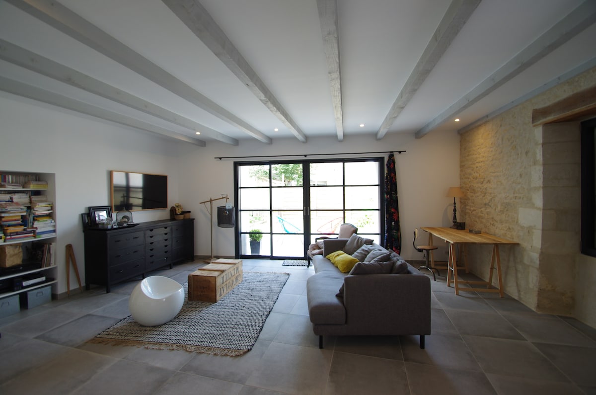 A spacious living area features sleek, contemporary furnishings and exposed wooden beams. A large window allows natural light to enter, illuminating a soft rug and minimalist décor. A dark cabinet holds books, while a rustic table and light armchair create a functional workspace.