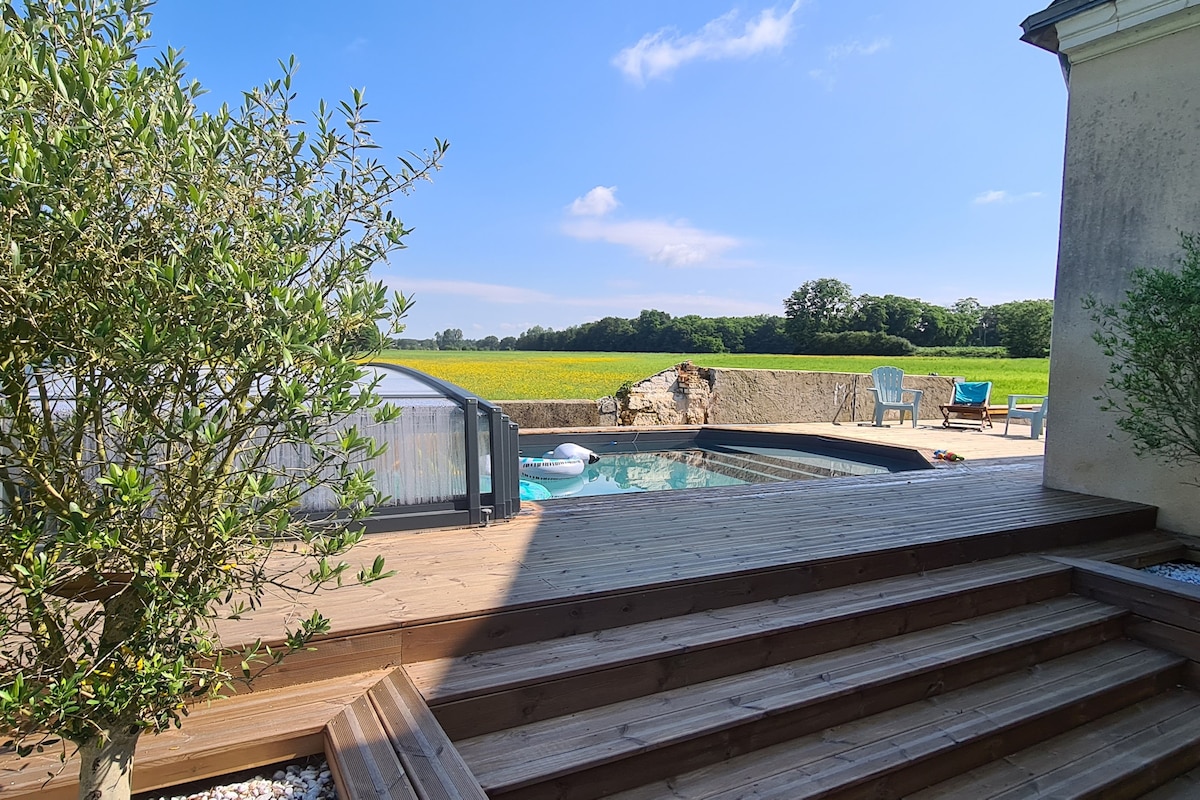 The outdoor area features a wooden deck surrounding a private pool, complemented by sun loungers and a view of expansive fields. Lush greenery, including small trees, frames the area, enhancing the serene atmosphere. The sky is clear and bright, creating a peaceful setting.