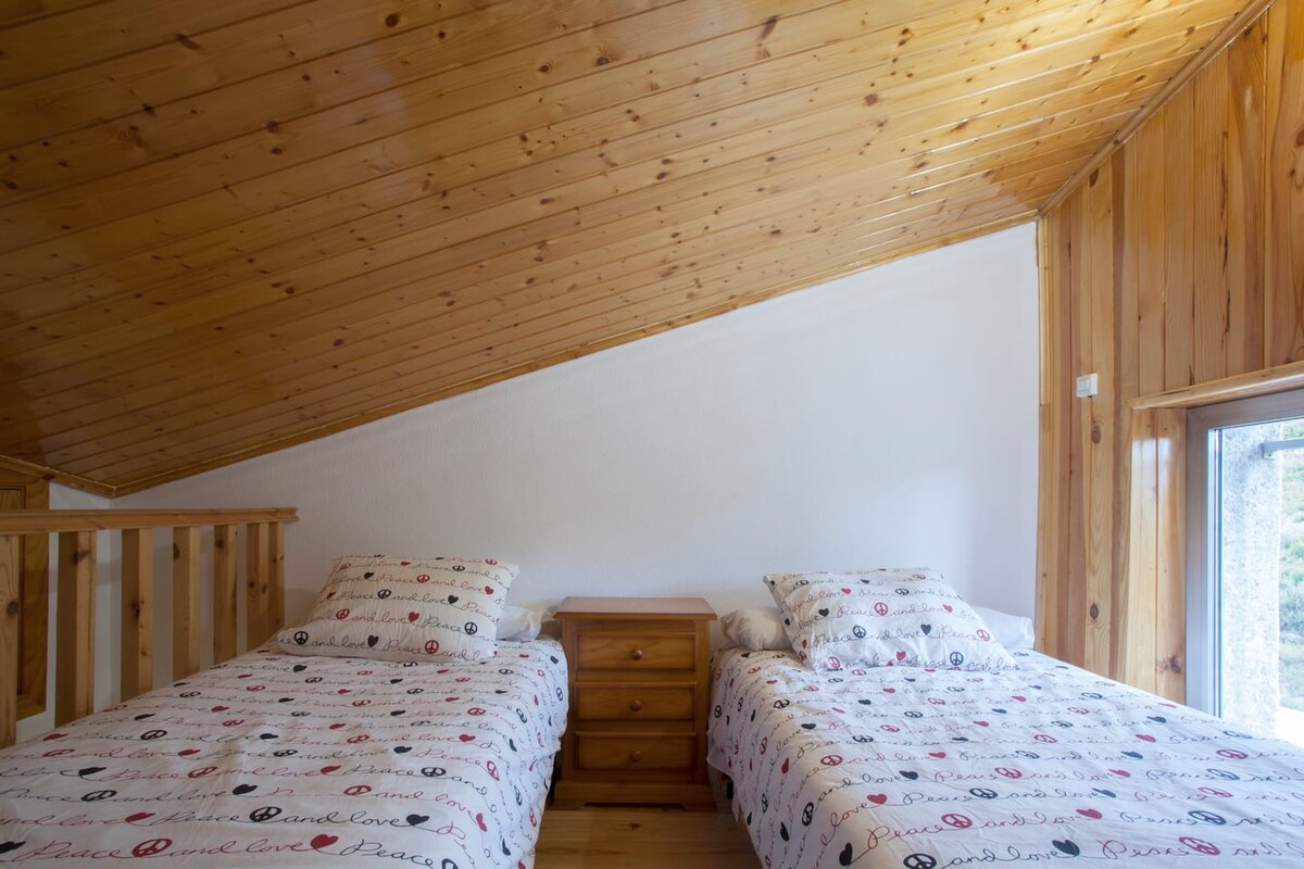 The sleeping area features two twin beds with patterned bedding, positioned beside a wooden nightstand. Natural light enters through a window, illuminating the wooden ceiling and warm-toned paneling that creates a cozy atmosphere.