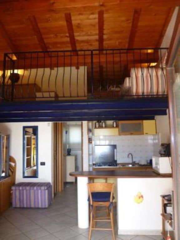 An interior view of the cozy mini-apartment features a loft area with seating, open to a fully equipped kitchenette and bar-style seating below. Natural light streams in from the window, highlighting the wooden ceiling and warm tones of the space.