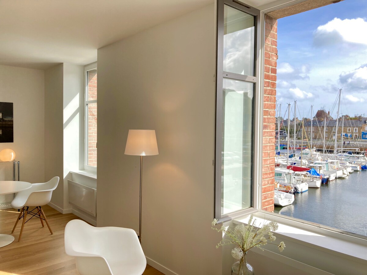 Large windows flood the contemporary living space with natural light, offering views of the marina. A sleek lamp stands beside a modern chair, while soft shadows create a serene atmosphere. The tranquil harbor scene, featuring numerous docked boats, is visible just outside.