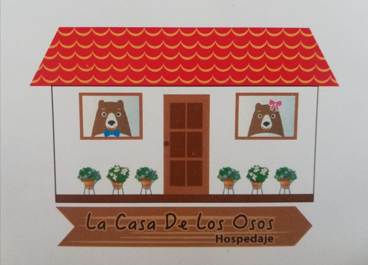 An illustrated house, named 'La Casa De Los Osos,' features a red-roofed structure with two windows displaying cartoon bears wearing a bow tie and a bow. Flowers are positioned below the windows, adding a cheerful touch. The name is presented on a wooden-style sign beneath the house.