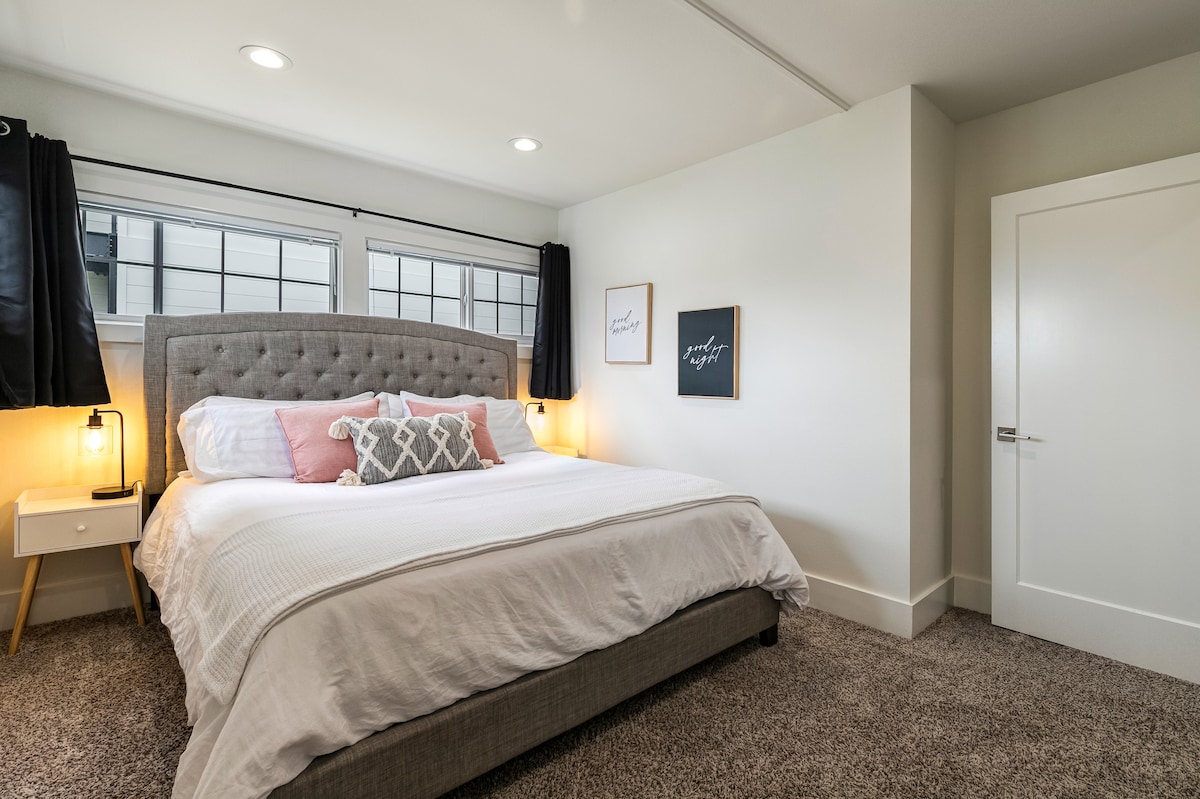 A master bedroom features a king-sized bed with soft linens and four pillows. Nightstands with lamps and USB ports are positioned on each side. Blackout curtains cover the windows, providing options for light control in the spacious room.
