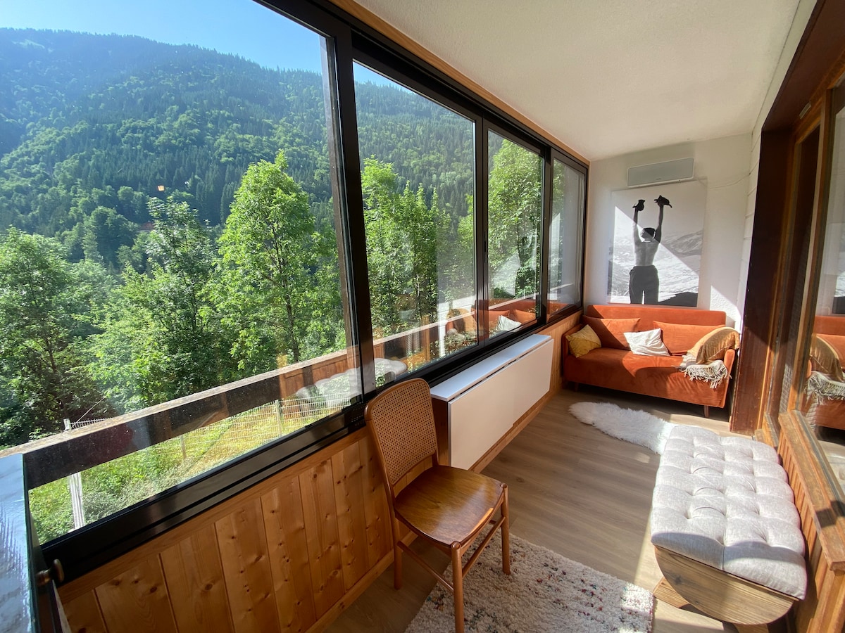 A bright verandah features large windows offering scenic views of the surrounding mountains and lush greenery. A cozy orange sofa and a beige tufted bench are positioned alongside a wooden chair. Soft light illuminates the space, enhancing the connection with nature.