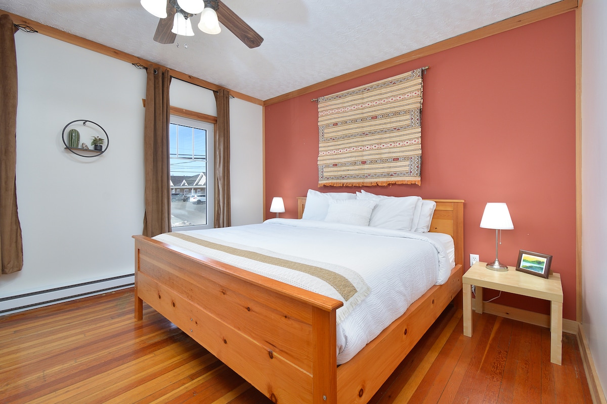 A spacious bedroom is furnished with a large wooden bed topped with white bedding. Two bedside lamps provide soft lighting, and a piece of textile art hangs above the headboard. Natural light enters through a window, while hardwood floors add warmth to the space.