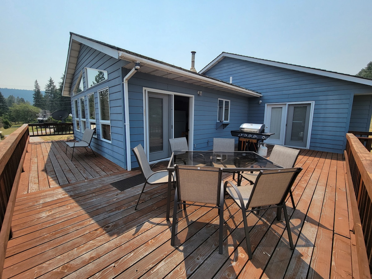 An outdoor deck features a set of six metal-framed chairs surrounding a glass-top table. The light blue exterior of the house is complemented by large windows, allowing natural light to fill the space. A barbecue grill is positioned nearby, enhancing outdoor dining options.