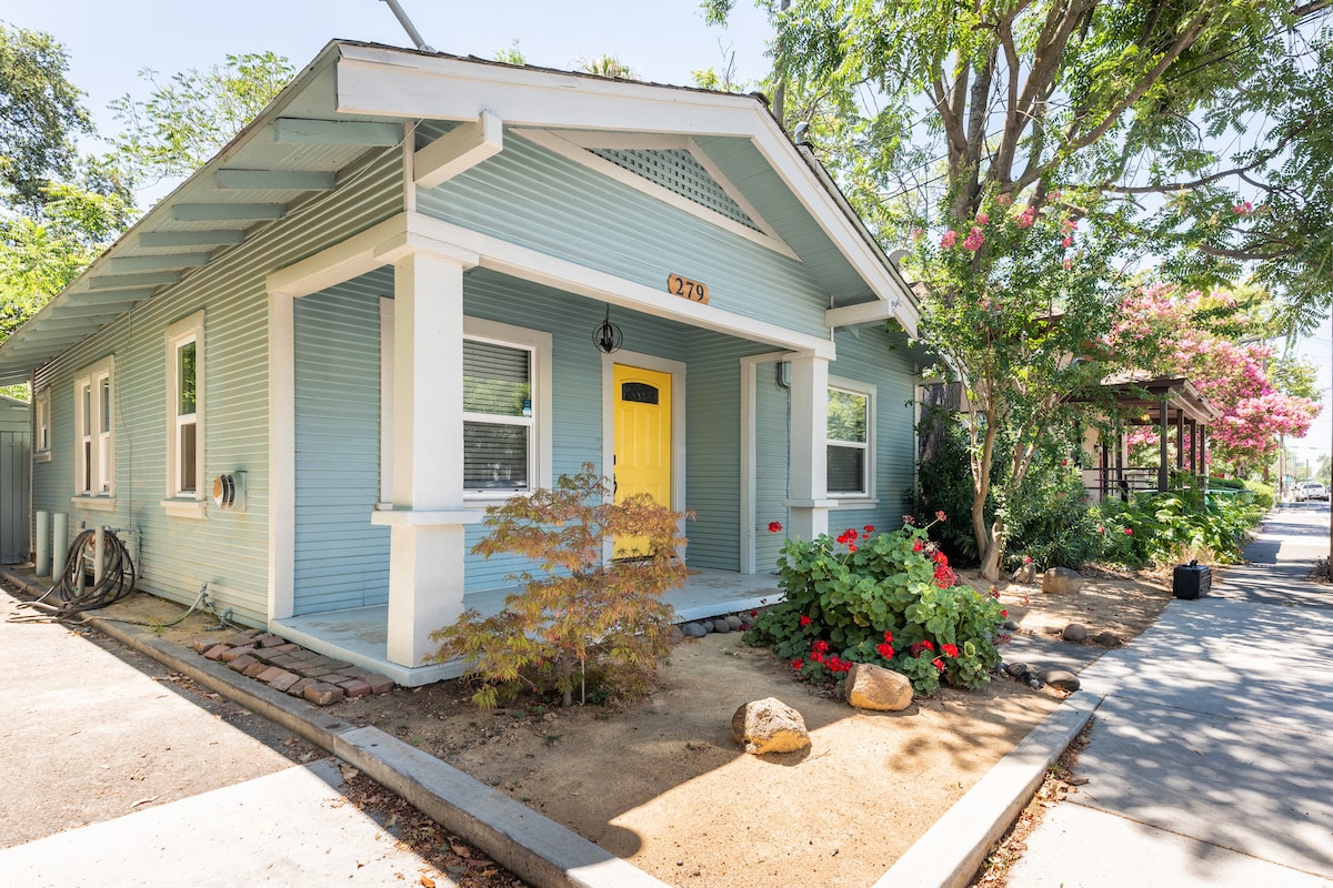 A charming blue house with a bright yellow front door is framed by well-maintained landscaping. It features a sandy garden area with flowering plants and rocks. Shade is provided by surrounding trees, and a pathway leads to the entrance.