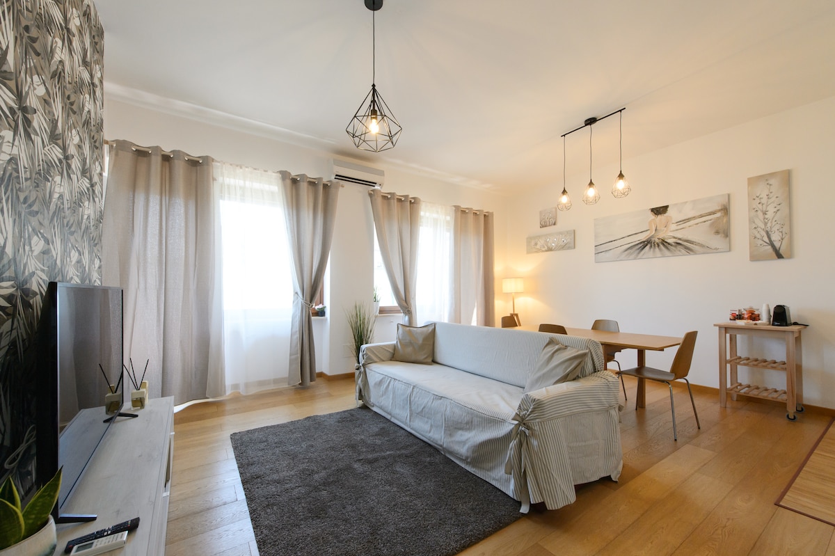 A well-lit living area features large windows draped with sheer curtains, allowing natural light to fill the space. A neutral-colored sofa occupies the center, accompanied by a dining table for four. Pendant lights hang from the ceiling, and a small service cart is positioned to one side.