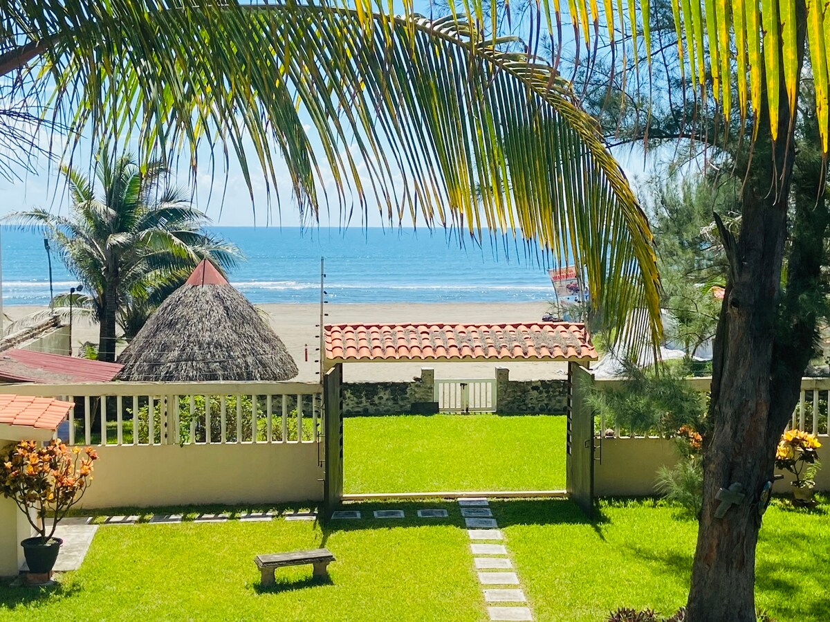 A well-maintained garden is framed by palm trees and a stone pathway, leading to a stunning view of the ocean. A traditional palapa roof can be seen in the distance, while gentle waves lap at the shore, creating a serene coastal atmosphere.