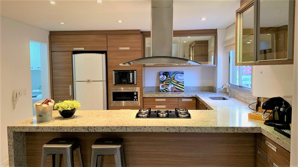 A modern kitchen is equipped with sleek cabinetry and a granite countertop. Stainless steel appliances, including a refrigerator, microwave, and oven, are visible. A central island features a gas cooktop and seating for two, complemented by natural light from large windows.