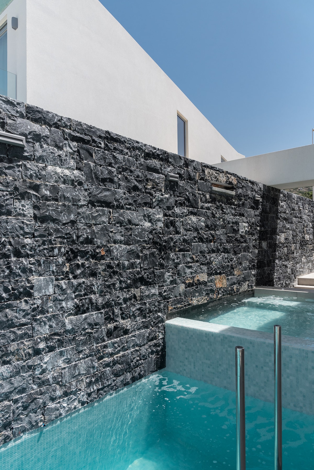 The image captures a sleek infinity pool adjacent to a textured stone wall, featuring a serene water feature. Sunlight reflects off the water's surface, enhancing the calm ambiance. A clear blue sky stretches above, creating a tranquil outdoor space.