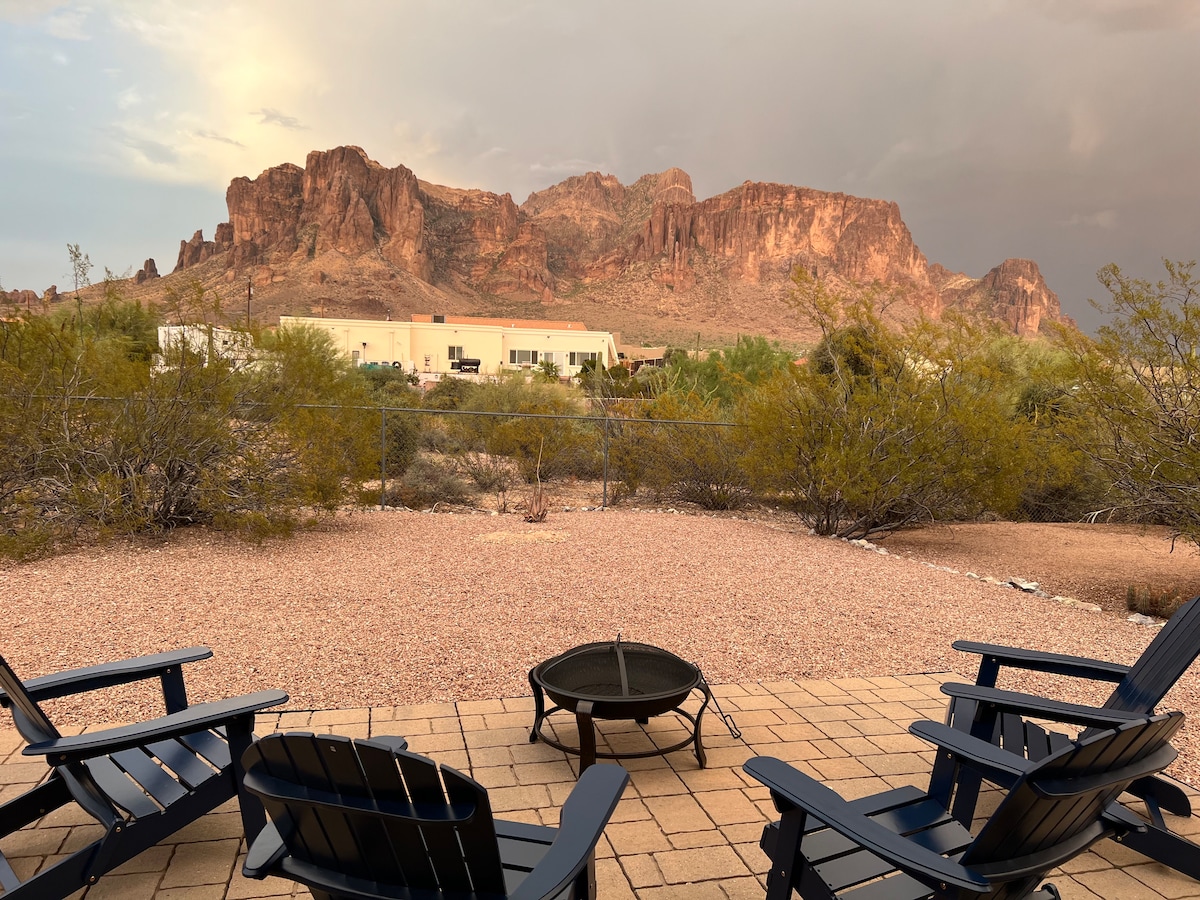 Four dark blue outdoor chairs surround a stone fire pit, overlooking a desert landscape. The distant Superstition Mountains rise majestically under a partly cloudy sky. Sparse vegetation includes low shrubs and cacti, providing a natural contrast to the stone patio.