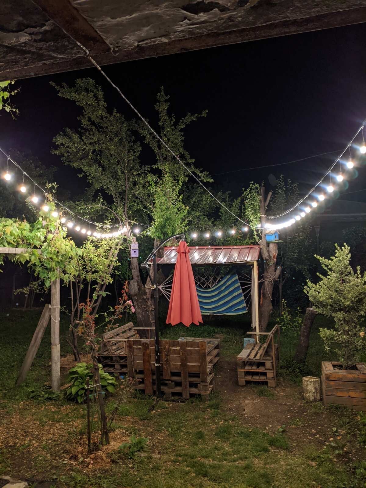An outdoor area is softly illuminated by string lights, creating a serene ambiance. A shaded seating space with a colorful umbrella is surrounded by greenery. Wooden pallets serve as rustic seating options, enhancing the space's natural charm.