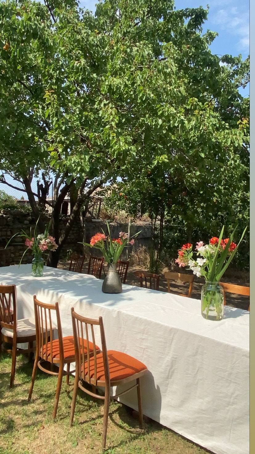 A long table is set under a lush tree, adorned with vases of fresh flowers. Wooden chairs with orange cushions surround the table, creating a welcoming space for gatherings. Sunlight filters through the leaves, enhancing the outdoor setting.