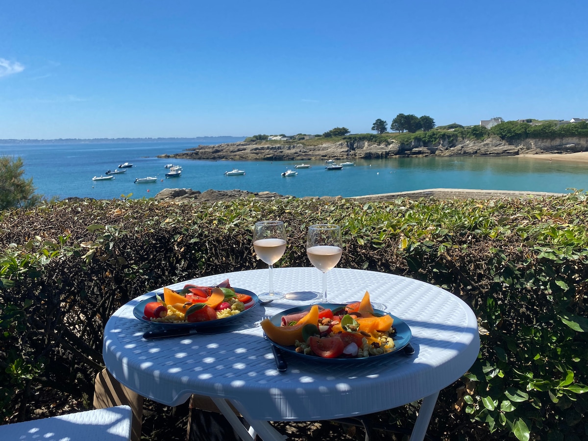 A table is set with two plates of fresh fruit and two glasses of rosé, overlooking a serene coastal view. Boats are anchored in the calm waters, with rocky shores and greenery visible in the background under a clear blue sky.