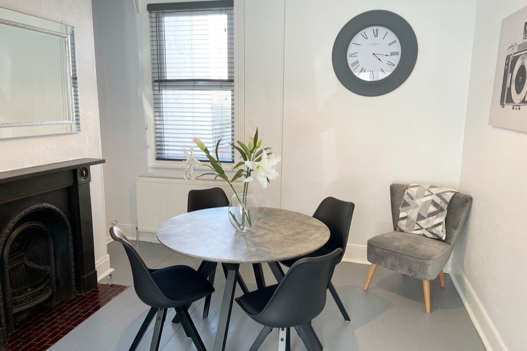 A dining area features a round table surrounded by four black chairs. A soft gray armchair is positioned nearby. Natural light enters through window coverings, and a clock is mounted on the wall. A decorative photo adds interest to the space.