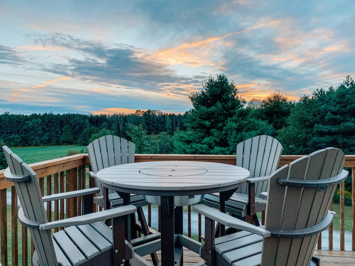 A round table surrounded by five Adirondack chairs rests on a wooden deck. The scene is complemented by a colorful sky at sunset above an expanse of trees, providing a peaceful outdoor setting.