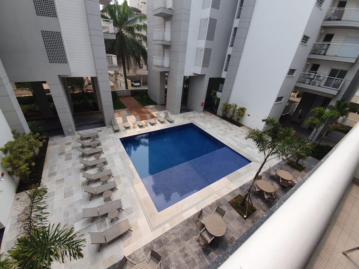 A serene pool area is framed by modern buildings, featuring a rectangular swimming pool with clear blue water. Sun loungers are arranged neatly around the pool, while stylish tables and chairs are positioned nearby, surrounded by landscaped greenery and palm trees.