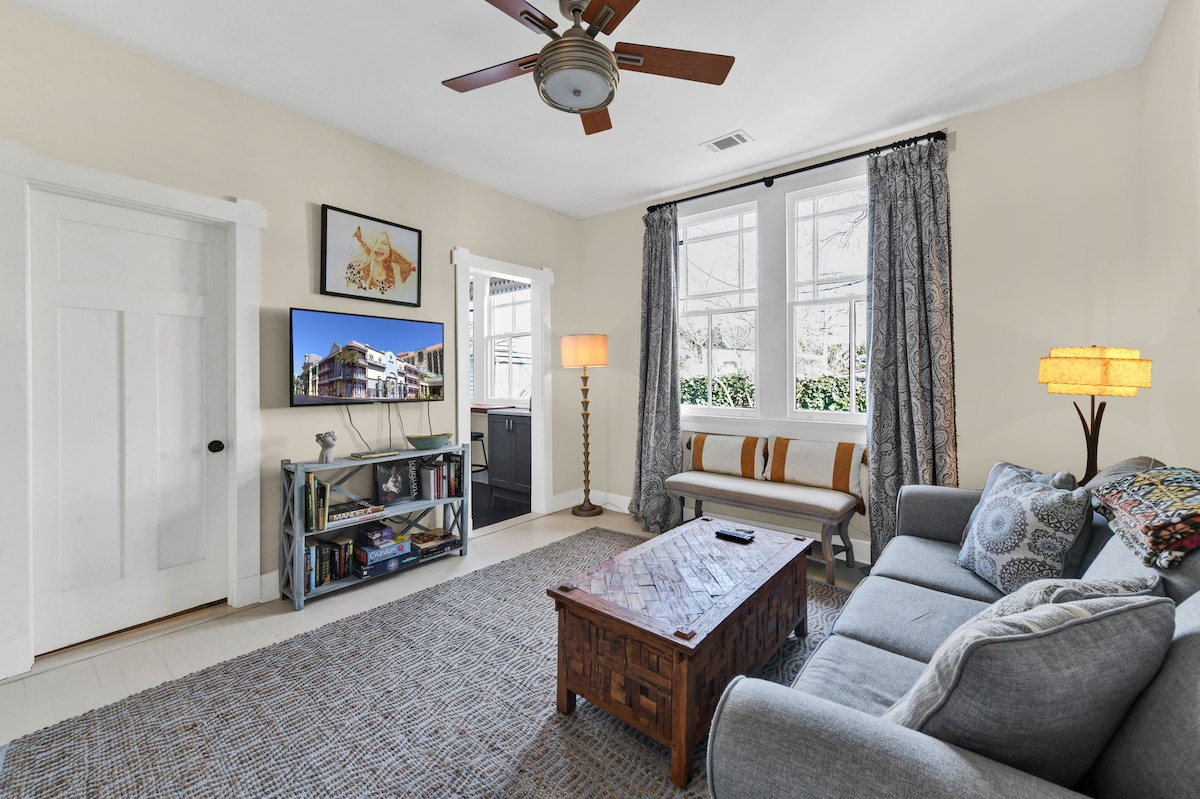 A comfortable living area features a gray sofa and a wooden coffee table on a textured rug. Two large windows allow natural light to brighten the space, complemented by a ceiling fan and a standing lamp. Decorative artwork is displayed on the wall.