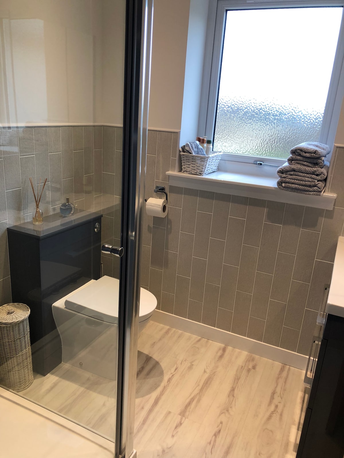 An ensuite bathroom is showcased with modern fixtures. A sleek toilet and a glass shower enclosure are present. Natural light enters through a frosted window. Neatly arranged towels and toiletries are placed on a shelf, contributing to a tidy and functional space.