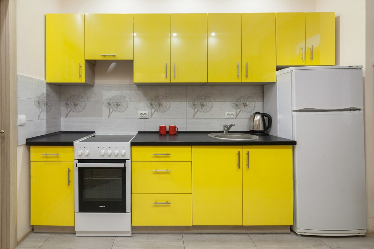 A modern kitchen features vibrant yellow cabinets and black countertops. Appliances include a refrigerator and an oven with a gas stovetop. A kettle and two red cups are positioned on the counter, enhancing the functional design of the space.