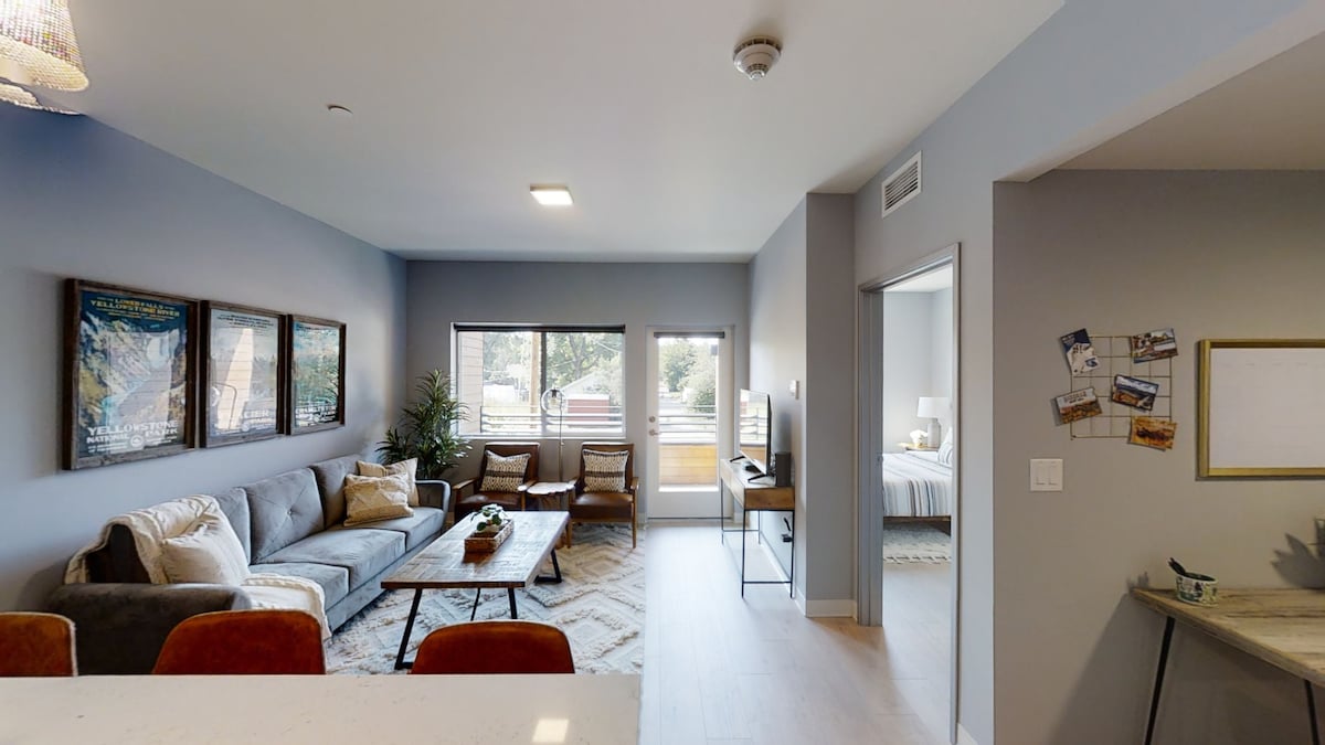 An open living space is featured, showcasing a comfortable gray sofa and two accent chairs. Art pieces are displayed on the wall, while natural light streams through large windows that lead to a private balcony. A door on the right provides access to the bedroom.