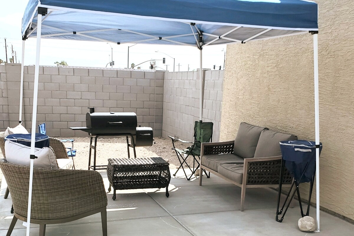 An outdoor patio area is sheltered by a blue canopy, providing shade over a seating arrangement that includes a couch and two chairs. A black grill is positioned on a table, and a small side table is visible. The space features a light-colored concrete floor.