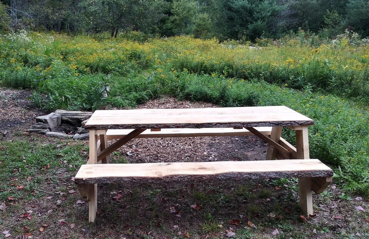 A rustic wooden picnic table with natural edges is set on a grassy area. Two attached benches flank each side. In the background, a diverse array of wildflowers and greenery creates a natural setting, complemented by a stone fireplace partially visible to one side.