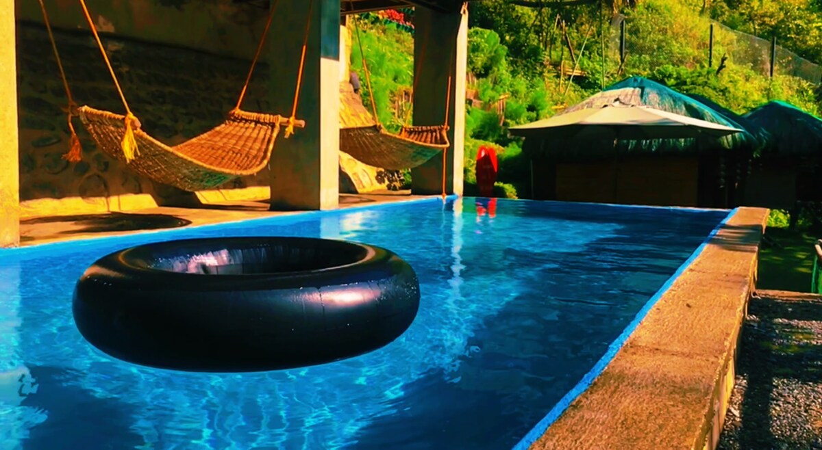 An inviting infinity pool is framed by stone walls, showcasing clear blue water. A black inflatable float rests on the surface. Two woven hammocks are suspended nearby, surrounded by lush greenery, offering a relaxed atmosphere to enjoy the outdoors.