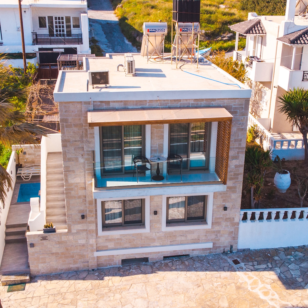 An exterior view of a two-story villa showcases a stone façade complemented by a spacious balcony. Two chairs and a small table are positioned on the balcony. The surrounding area features greenery and stone pathways, enhancing the villa's charming appeal.