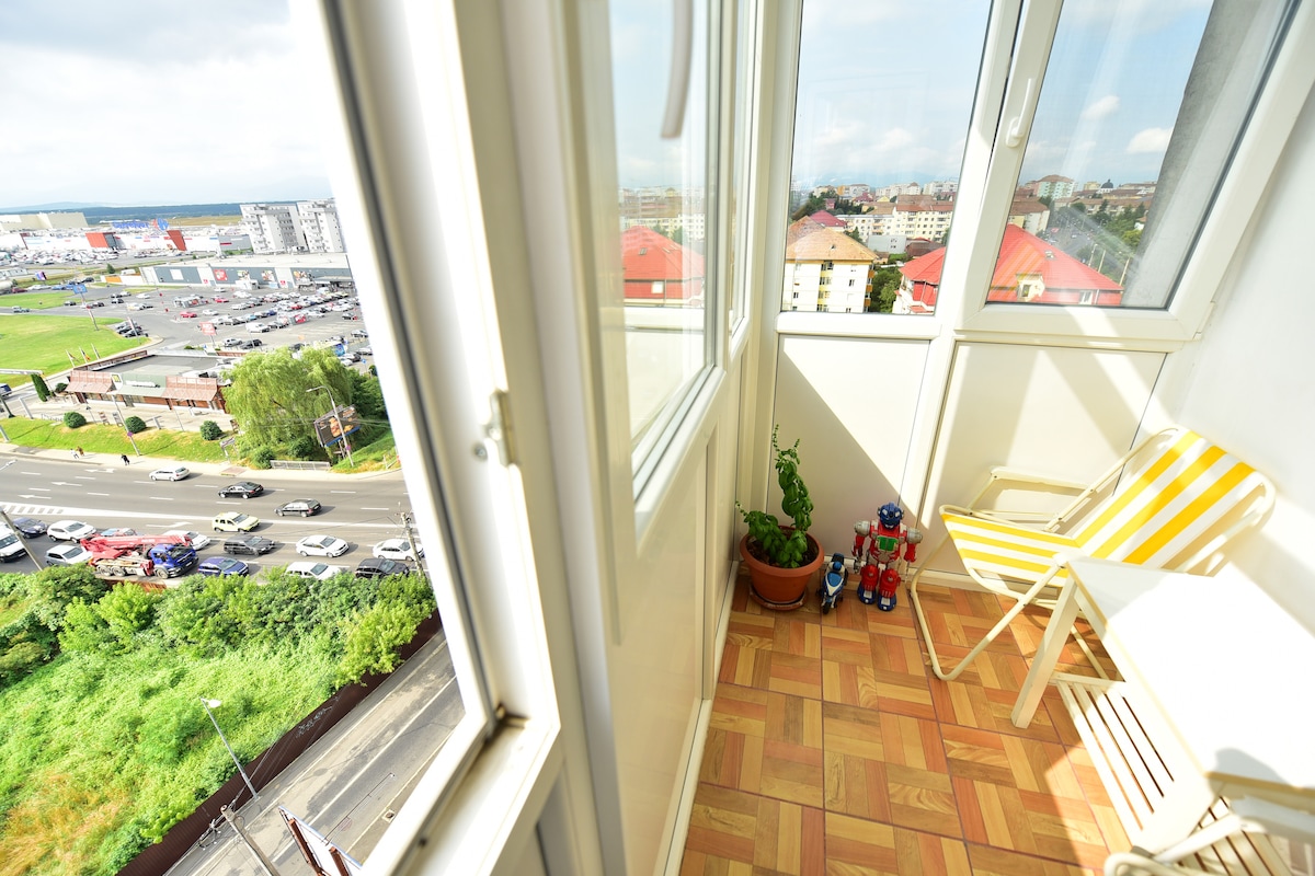 A bright sunroom is enclosed by large glass windows, providing expansive views of the surrounding area. A potted plant sits next to colorful toys, and a yellow striped chair is positioned near a light-colored wooden table on the tiled floor.