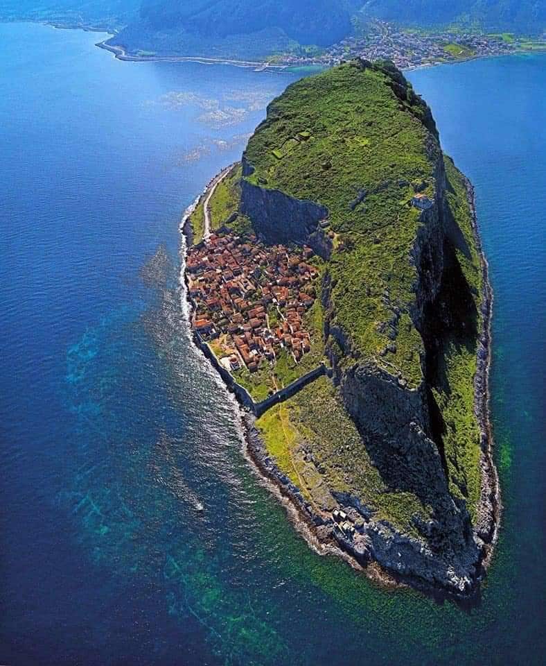 An aerial view captures the island of Monemvasia, showcasing its rugged, green cliffs rising from the clear blue waters. The medieval fortress appears nestled among the cluster of buildings, reflecting the traditional architectural style harmoniously set against the natural landscape.